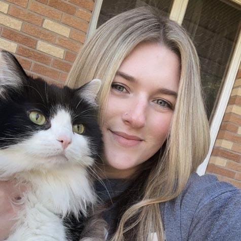 A woman with long blonde hair holds a black and white cat while standing outside in front of a brick wall and window.