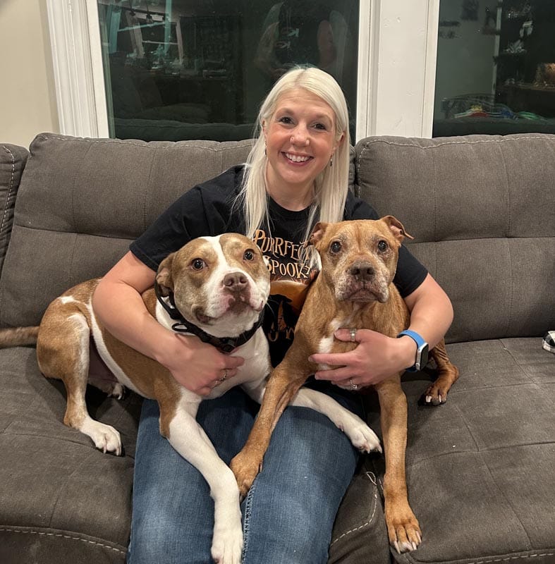 A woman with long blonde hair sits on a gray couch, smiling, with two brown and white dogs, one on each side of her.