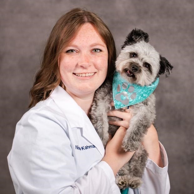 A woman in a white lab coat holds a small gray dog wearing a teal bandana, both facing the camera against a plain gray background.
