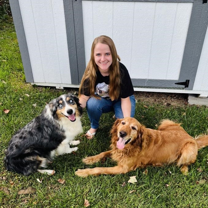 A woman kneels on grass beside a black, white, and gray dog sitting and a golden retriever lying down, all in front of a white shed.