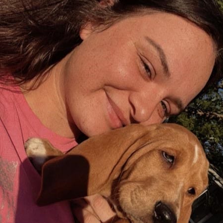 A woman with brown hair smiles while holding a brown puppy with long ears outdoors in sunlight.