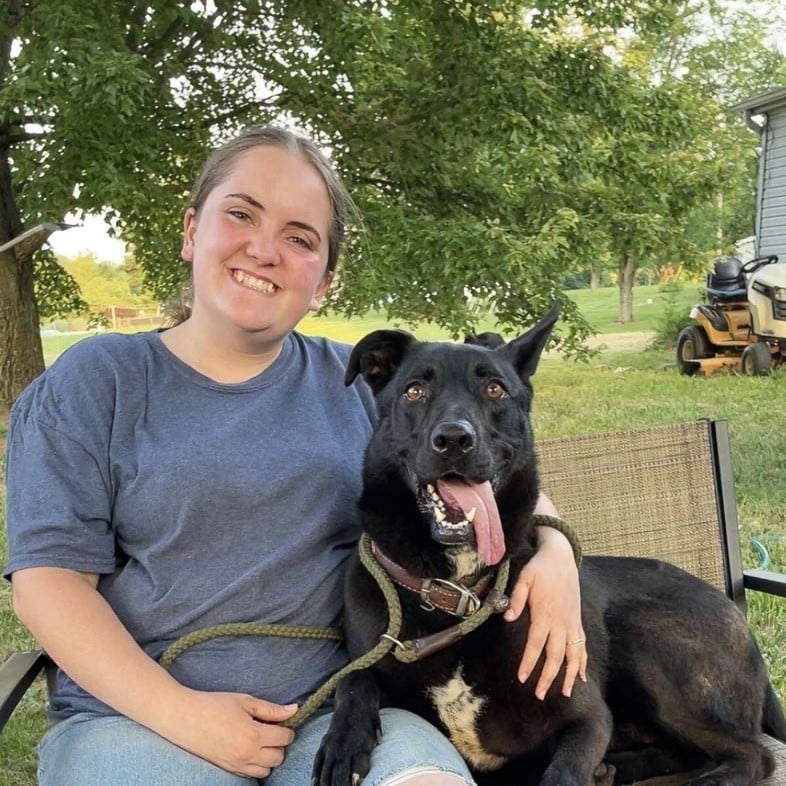 A woman sits outdoors on a chair, smiling with her arm around a large black dog whose tongue is out. Trees, grass, and a riding lawn mower are visible in the background.