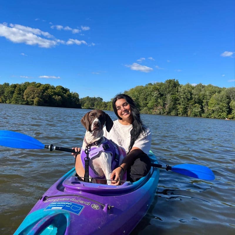 A woman and a dog sit together in a blue kayak on a calm lake, surrounded by trees under a clear blue sky.