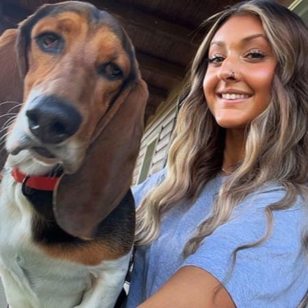 A woman with long wavy hair and a nose ring smiles next to a brown and black dog with long ears, both posing for a selfie outdoors.