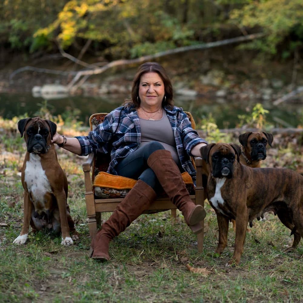 A woman sits in an armchair outdoors with two boxers standing on her left and one boxer standing on her right, near a wooded area and a creek.