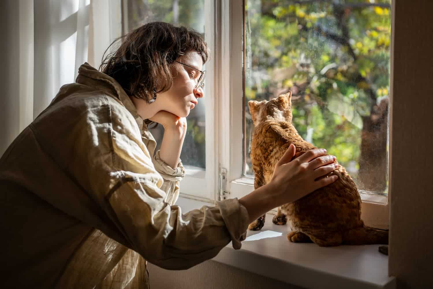 A person with glasses sits by a window, gently petting a cat as both look outside at the sunlit greenery.